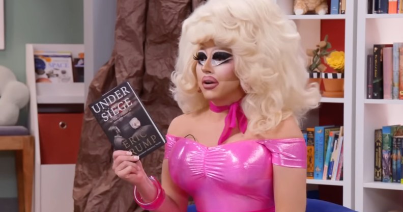 Trixie Mattel in her classic pink dress and big blonde wig holding Eric Trump's book in a school library.