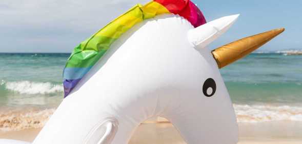 White inflatable unicorn float with rainbow mane and gold horn rests on a sandy beach with ocean waves in the background