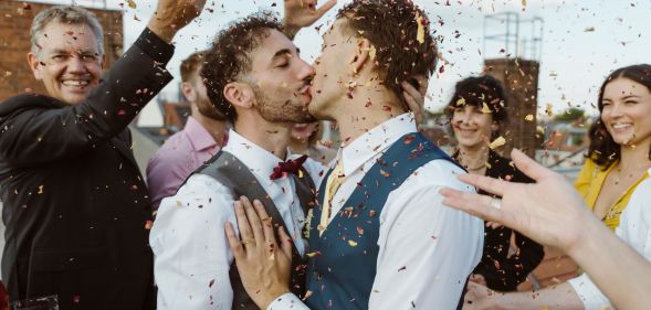 Two men kiss surrounded by a crowd of people throwing confetti.