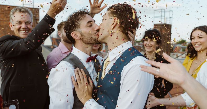 Two men kiss surrounded by a crowd of people throwing confetti.