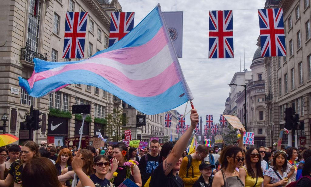 A crowd of people, one of which is holding a trans Pride flag, stand in a London city with Union Jack banners across the streets.