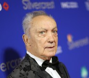 Udo Kier wearing a white shirt and black suit, on a red carpet standing against a blue background