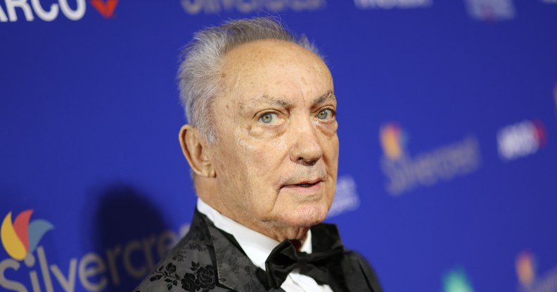 Udo Kier wearing a white shirt and black suit, on a red carpet standing against a blue background