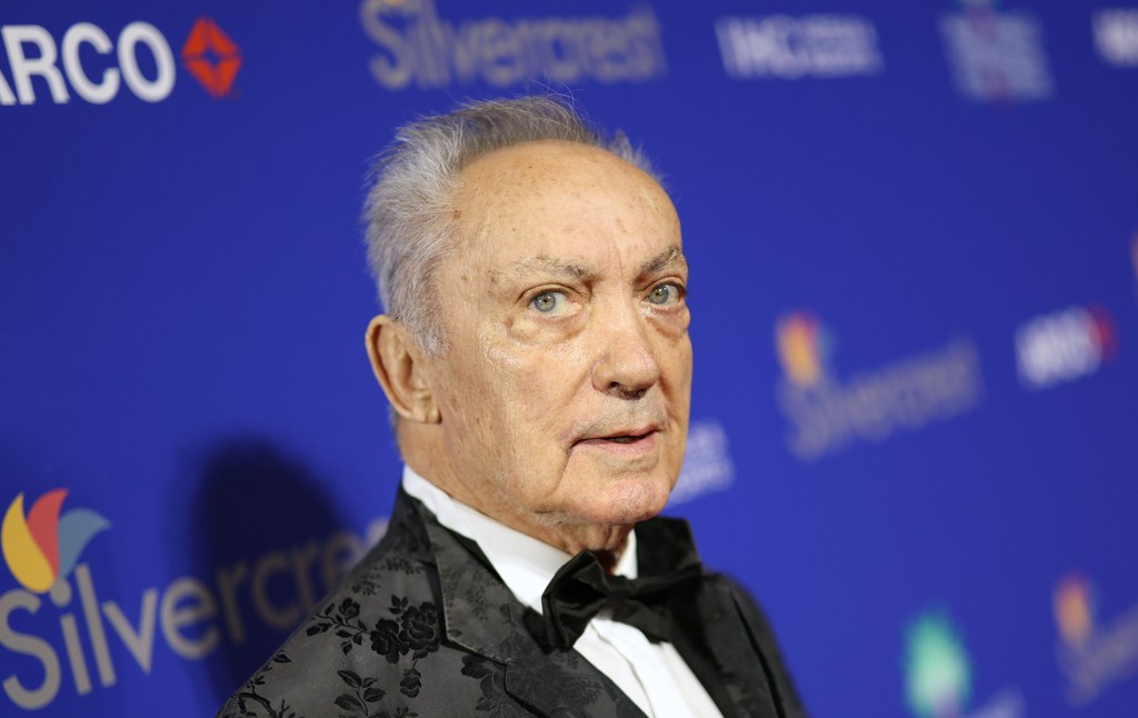 Udo Kier wearing a white shirt and black suit, on a red carpet standing against a blue background