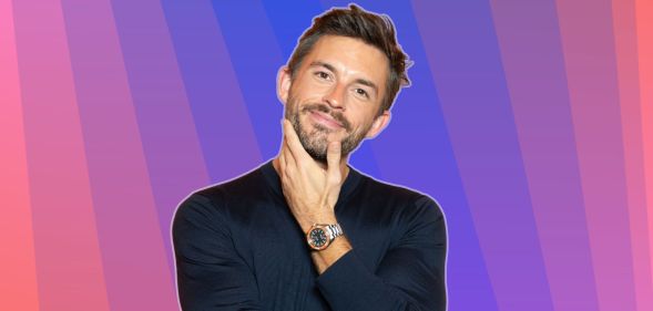 Jonathan Bailey smiles and places his hand on his chin. He is against a multi-coloured background.