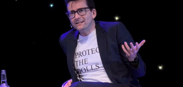 David Tennant on stage wearing a Protect The Dolls white t-shirt under a suit jacket