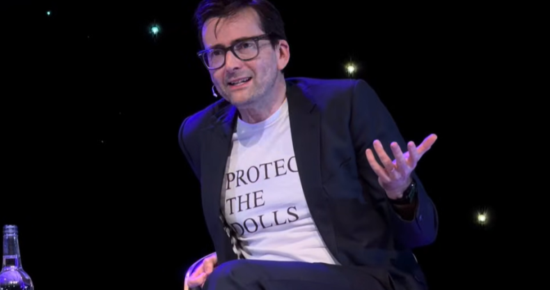 David Tennant on stage wearing a Protect The Dolls white t-shirt under a suit jacket
