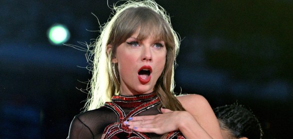 Taylor Swift performs onstage during the Eras tour. She's looking shocked with her hand on her chest.