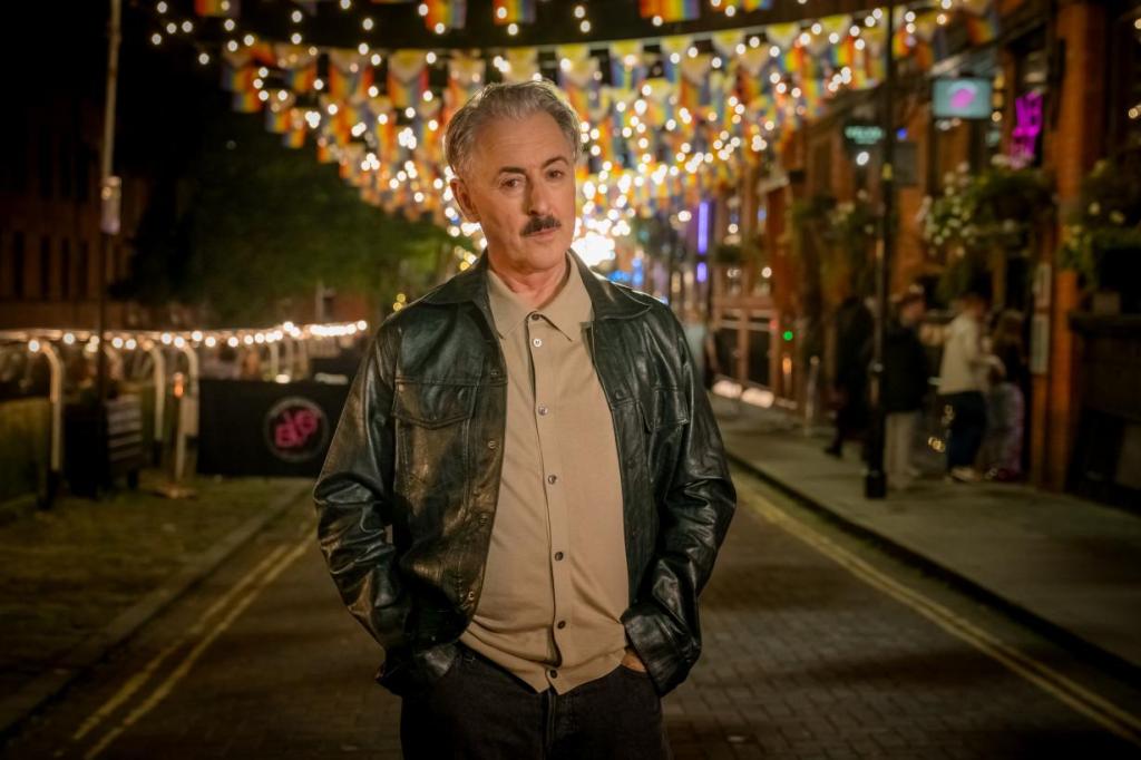 Alan Cumming as Leo standing on a street with gay bars behind him at night, Pride flags and ligths