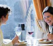 A queer couple has a meal at a fine dining restaurant. One person faces the camera and laughs at the other, who sits on the left away from camera smiling. On the table is food and wine. The person laughing holds a fork in hand and wears a white vest. The other has a black vest.