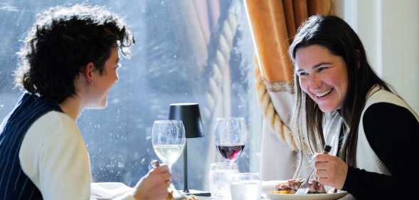 A queer couple has a meal at a fine dining restaurant. One person faces the camera and laughs at the other, who sits on the left away from camera smiling. On the table is food and wine. The person laughing holds a fork in hand and wears a white vest. The other has a black vest.
