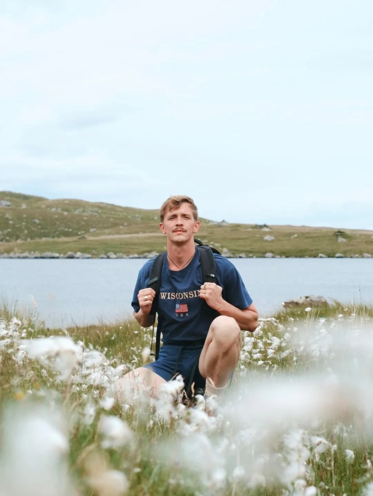 Stephen kneeling down amongst a field of white flowers, a lake behind him. Wearing a dark navy t shirt and shorts