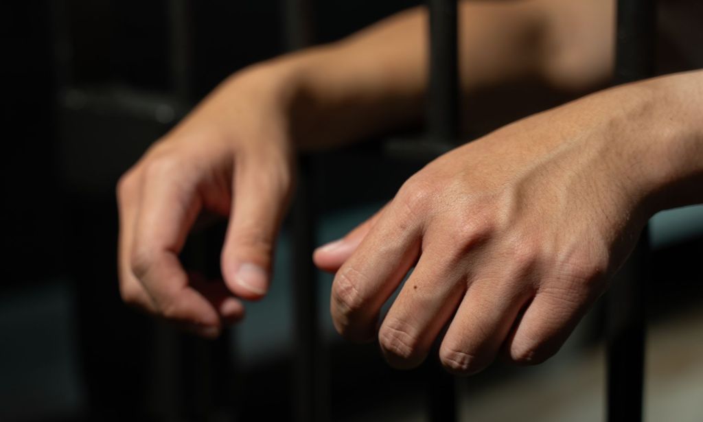 An unpictured person's hands leaning against prison bars.