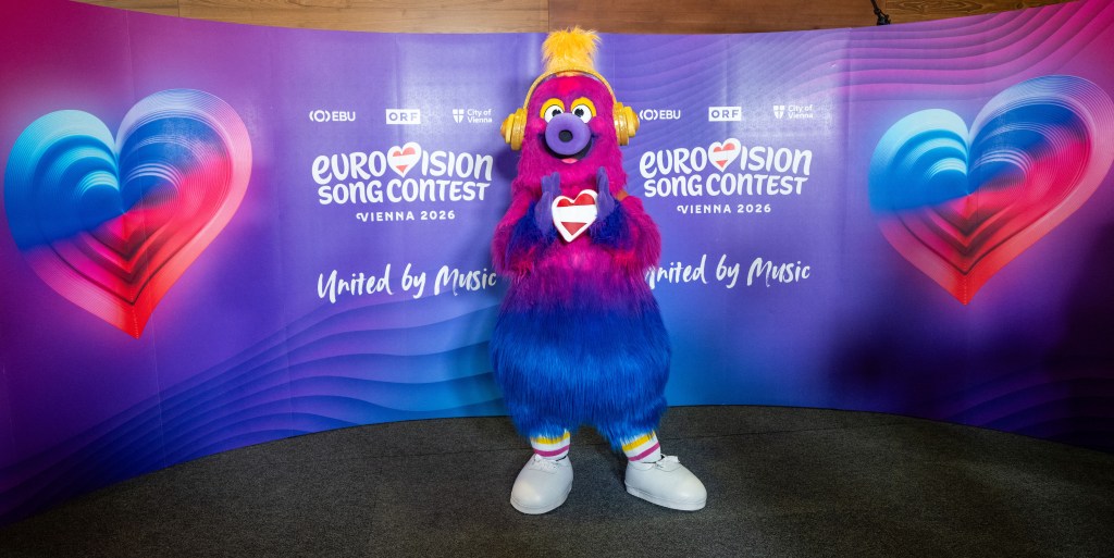 Auri is the mascot of the 2026 Eurovision Song Contest. A fluffy creature which has a pink head that fades to purple and then blue legs. Auri is wearing trainers and has yellow headphones on. Holiday Austria flag heart