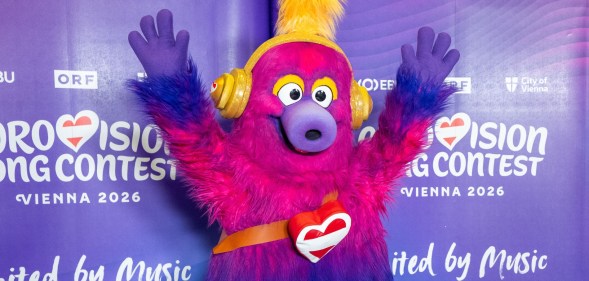Auri is the mascot of the 2026 Eurovision Song Contest. A fluffy creature which has a pink head that fades to purple and then blue legs. Auri is wearing trainers and has yellow headphones on. Holiday Austria flag heart