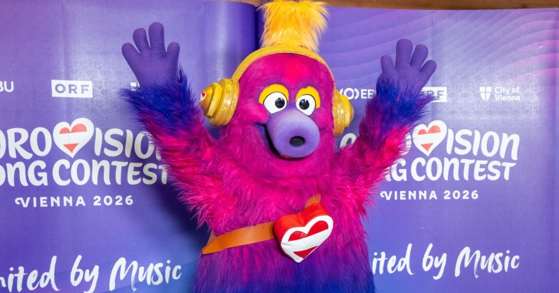 Auri is the mascot of the 2026 Eurovision Song Contest. A fluffy creature which has a pink head that fades to purple and then blue legs. Auri is wearing trainers and has yellow headphones on. Holiday Austria flag heart