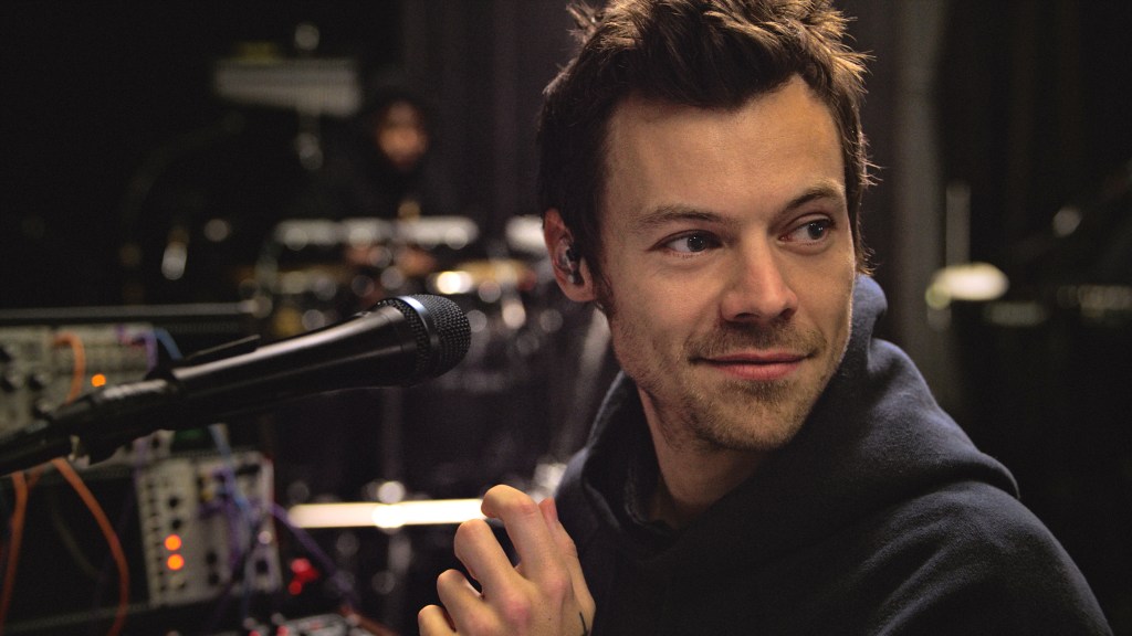 Harry Styles looking off screen in a rehersal room, he's wearing a dark navy hoodie.