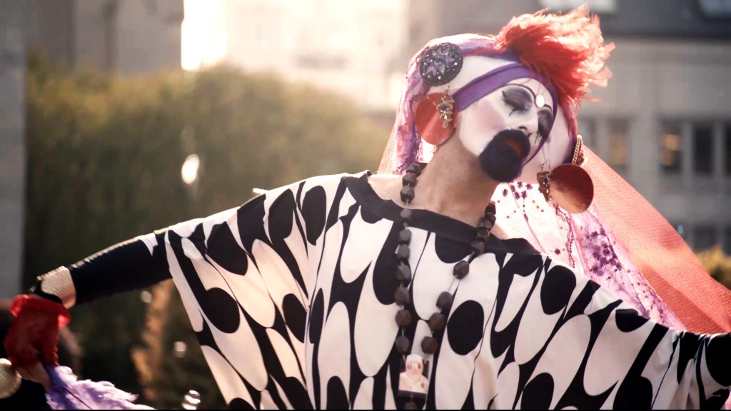Hunky Jesus still: a person dressed in drag with a black and white long dress and a purple and orange headpiece.