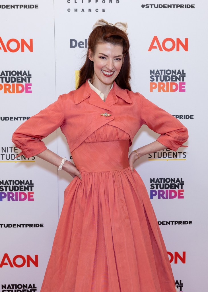 Jessica Kellgren-Fozard in a peach pink dress with a golden broach in the middle of her chest. Smiling at the camera.