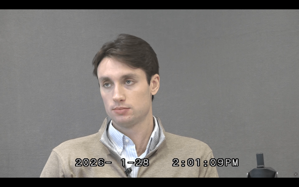 Justin Fox's testimony: he's sat against a grey background. Beige sip jacket over a blue and shite striped shirt.