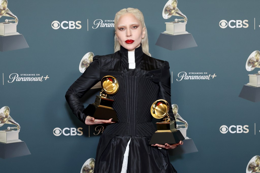 Lady Gaga standing with two Grammys in a long black dress with white details under.