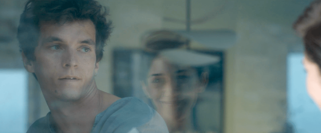 Fionn Whitehead as Philip in Satisfaction. He's wearing a blue toned top, looking through the window at a woman