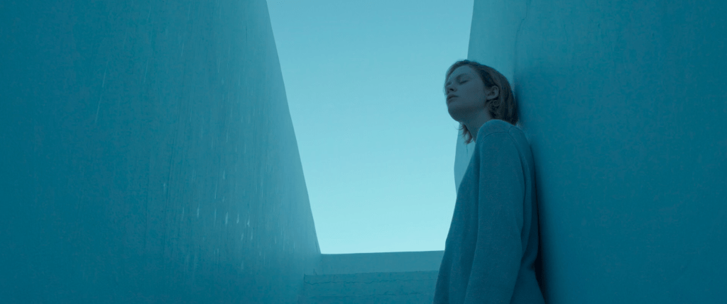 Emma Laird as Lola in Satisfaction. She's walking up pale white steps towards blue sky, the imaage a washed out blue tone