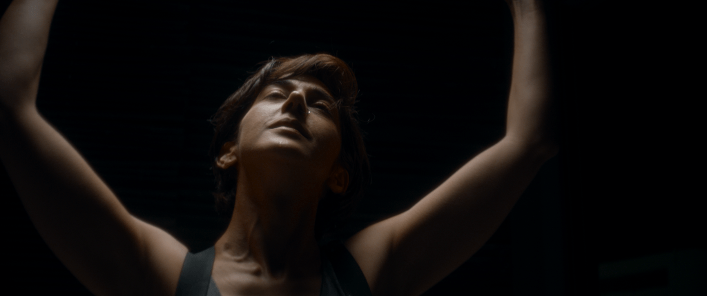 Amir Ebrahimi as Elena in Satisfaction. She's got her arms raised, dancing, against a black background.