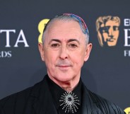 Alan Cumming attends the 2026 EE BAFTA Film Awards at The Royal Festival Hall. He's in a black suit jacket with pink and blue hair accessories. A silver brooche on his tie knot
