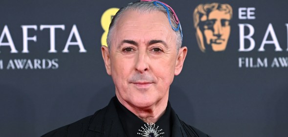 Alan Cumming attends the 2026 EE BAFTA Film Awards at The Royal Festival Hall. He's in a black suit jacket with pink and blue hair accessories. A silver brooche on his tie knot