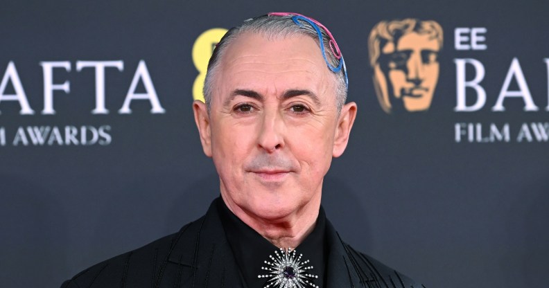 Alan Cumming attends the 2026 EE BAFTA Film Awards at The Royal Festival Hall. He's in a black suit jacket with pink and blue hair accessories. A silver brooche on his tie knot