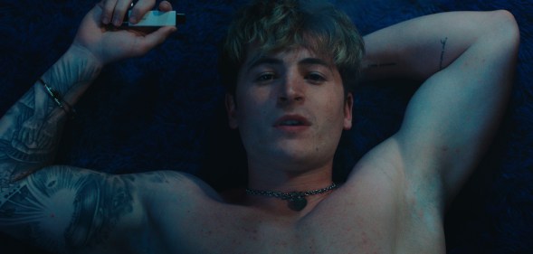 Kieron Moore stars as Aaron. He's topless, lying down and looking up at the camera with a vape in his hand.