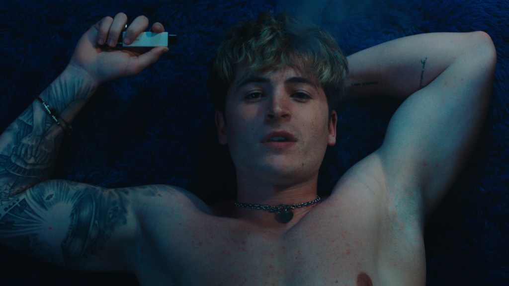 Kieron Moore stars as Aaron. He's topless, lying down and looking up at the camera with a vape in his hand.