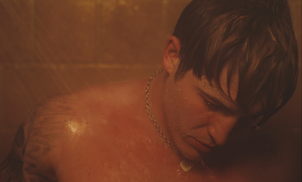 Aaron in the shower, water cascading on his shoulder. Image is orange-toned