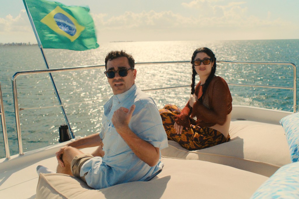 Dan Levy and Taylor Ortega star in Big Mistakes. The pair are sat on a sofa at the back of a boat