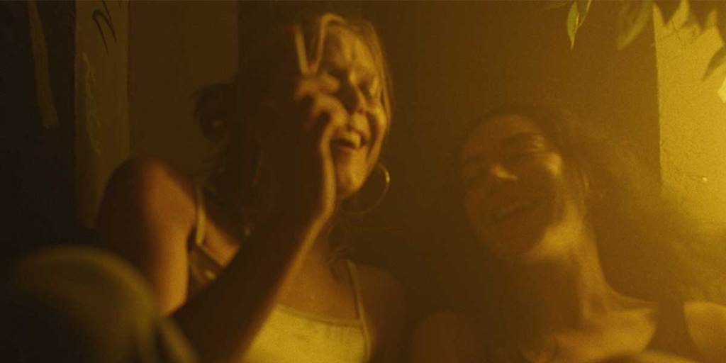 Erupcja still: Charli XCX and Lena Góra laughing and smoking in the film under yellow light.
