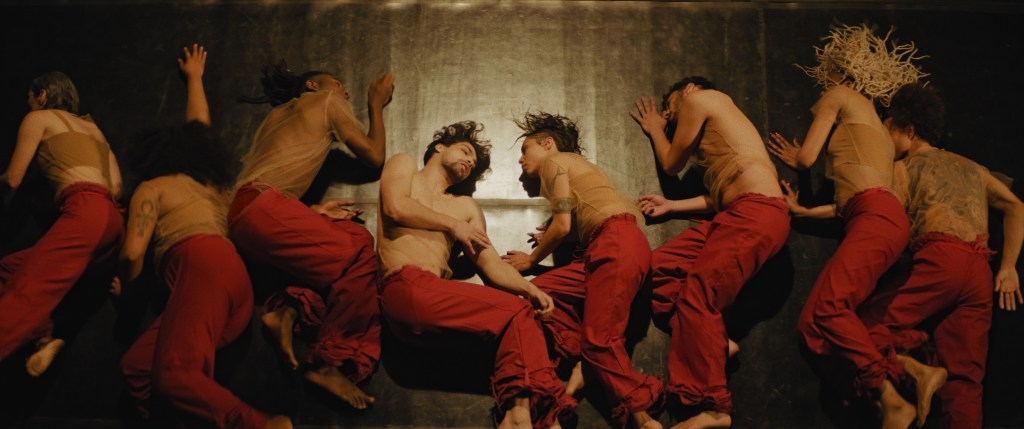 8 performers lying on a wooden floor in nude mesh tops and red trousers
