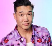 Joel Kim Booster in a jacket with purple faces and red lips. The background is hazy pink and blue.