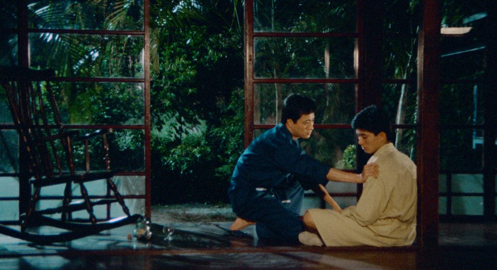 The Outsiders still, a man reaching out on another man's shoulder in a greenhouse-like building