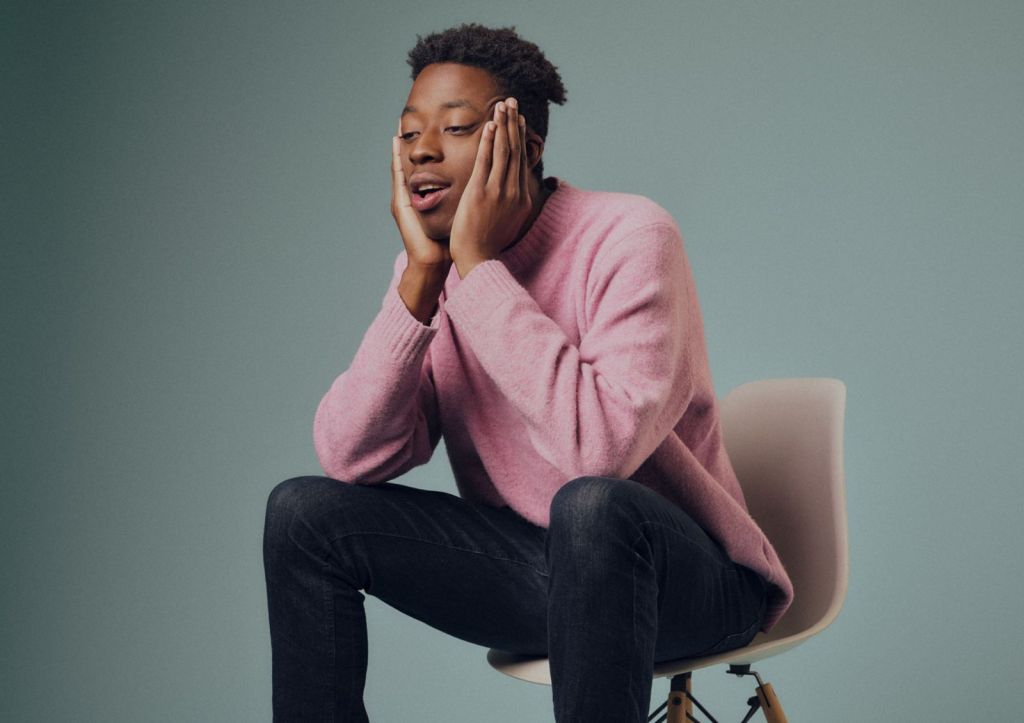 Ayo Adenekan: Homeland Insecurity. Ayo in a pink jumper sat on a small chair