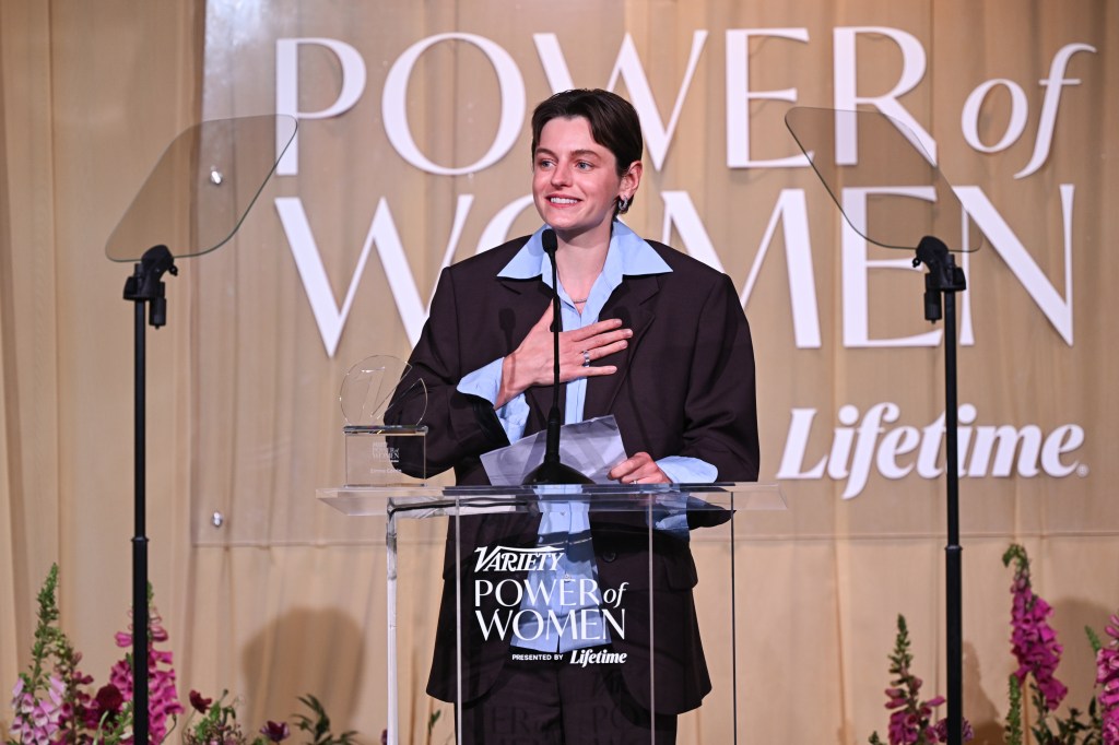 Emma Corrin at Variety 2026 Power of Women London. They're wearing a pale blue shirt under a brown suit, they're stood at a podium reading their speech