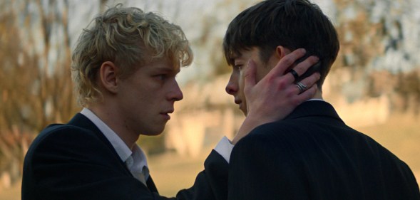 Leviticus still: two teen boys, the blonde cradling the cheek of the brunet, both in suits.