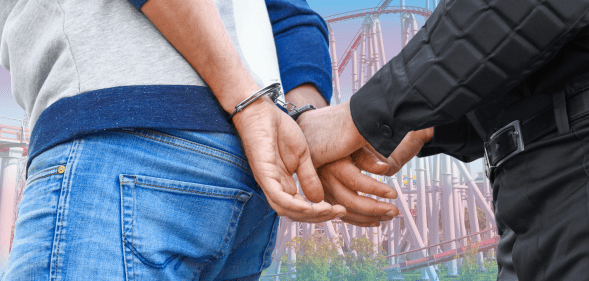 A man's hands in a pair of handcuffs as he is led away by police. The background shows a roller coaster with the trans flag colours overlaid.