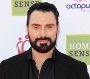 Rylan Clark smiles on a red carpet wearing a black shirt.