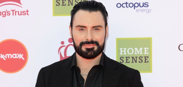 Rylan Clark smiles on a red carpet wearing a black shirt.