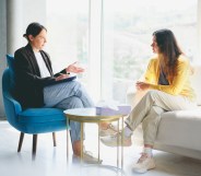 Two women are seated in a bright room with large windows. One woman, wearing a yellow sweater, is listening attentively while the other woman takes notes. The setting is professional, suggesting a counseling or meeting scenario. The room features a blue chair, a white sofa, and a small table with a tissue box. No visible copy space.