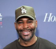 Karamo Brown smiling on a red carpet against a blue background. He's wearing a green LA cap and a black t-shirt
