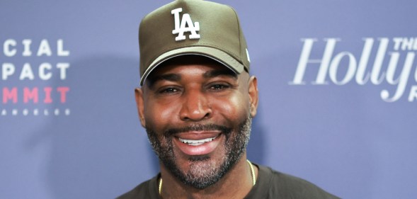 Karamo Brown smiling on a red carpet against a blue background. He's wearing a green LA cap and a black t-shirt