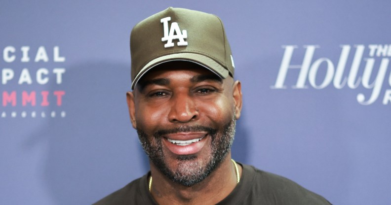 Karamo Brown smiling on a red carpet against a blue background. He's wearing a green LA cap and a black t-shirt