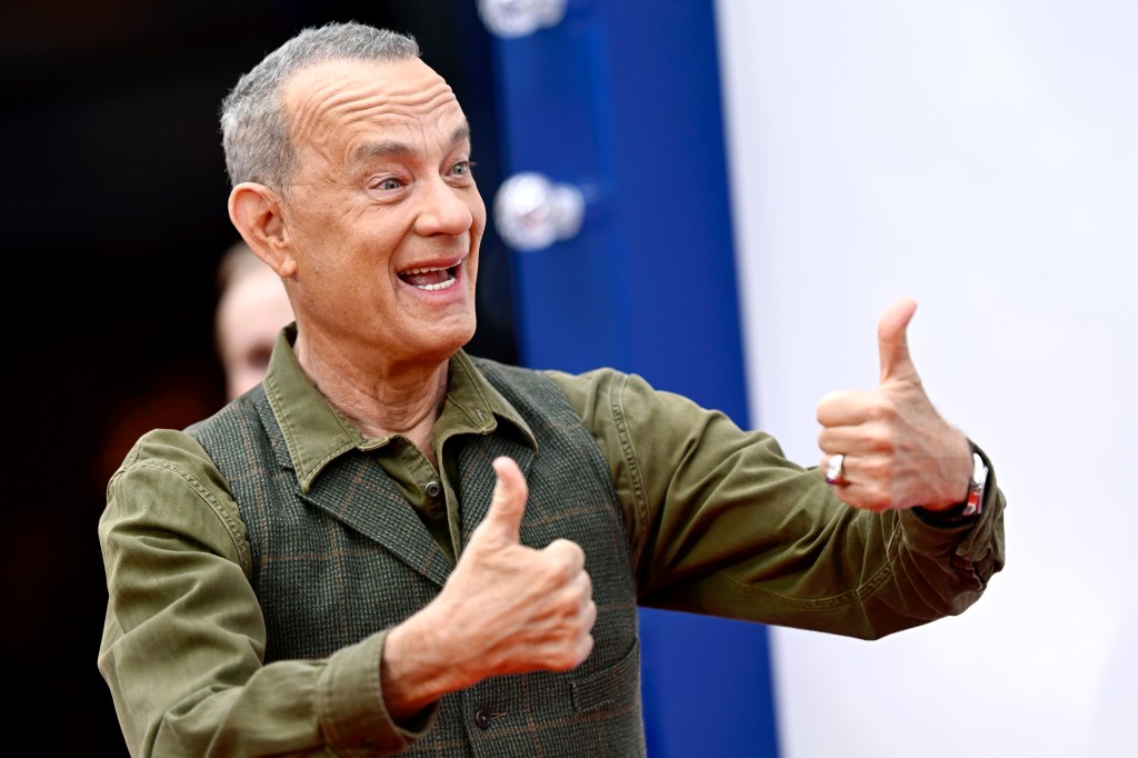 Tom Hanks on a red carpet in a green shirt, holding up two thumbs up.